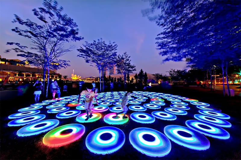 Children play on glowing, round platforms in a park at night. Buildings are visible in the background.